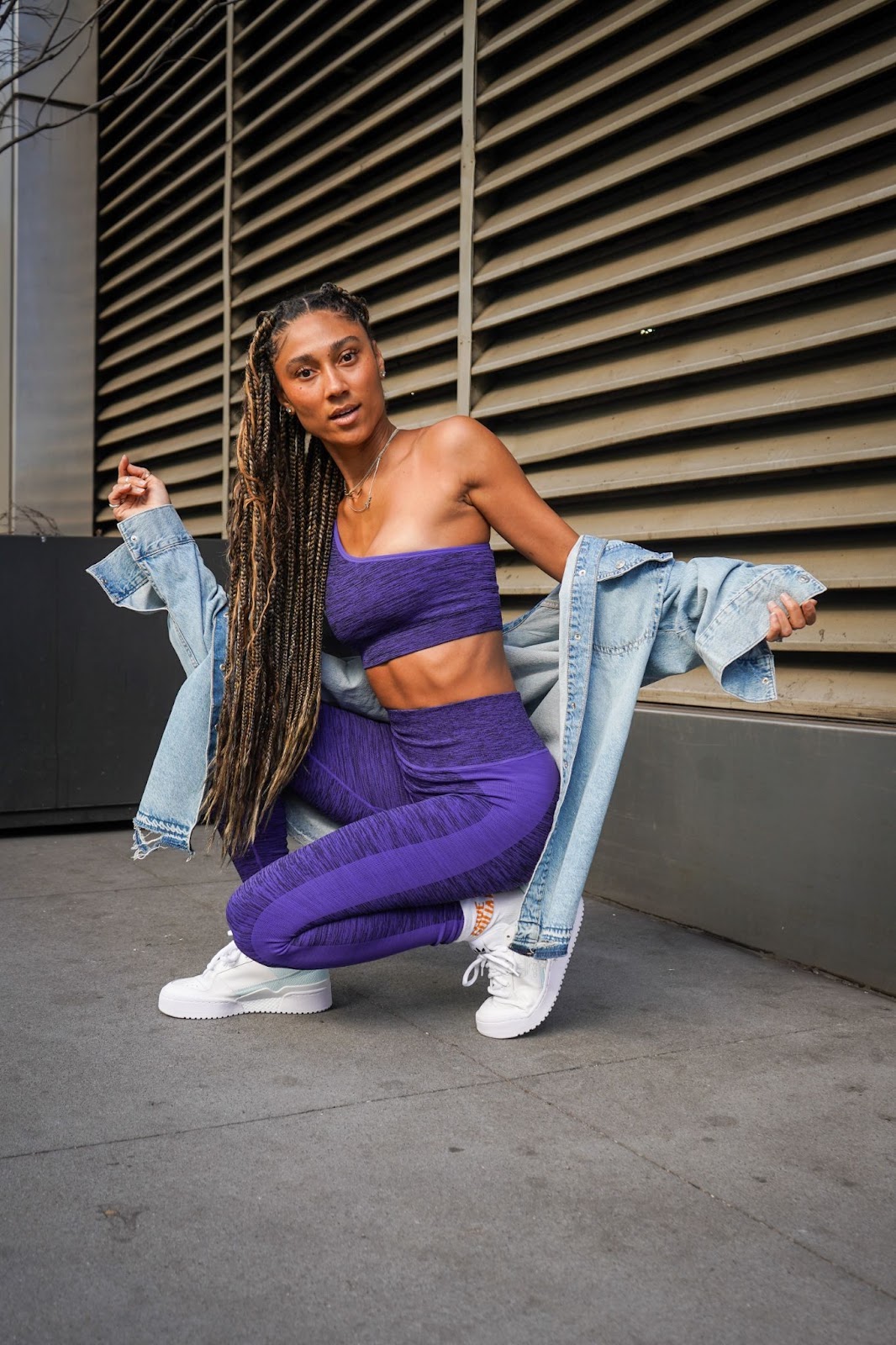 A woman in matching purple leggings and a sports bra, crouching down, and wearing an oversized denim sheet slung off of her shoulders.